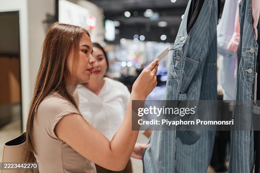 Asian women shopping for denim overalls in a clothing store. One examines the price tag closely. The scene captures a candid shopping decision in a modern retail environment. Asian women shopping for denim overalls in a clothing store. One examines the price tag closely. The scene captures a candid shopping decision in a modern retail environment.