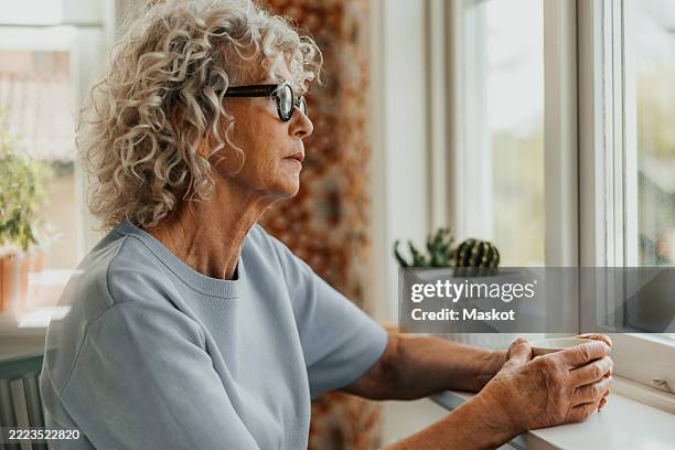 thoughtful senior woman holding coffee cup while looking through window at home - ensamhet bildbanksfoton och bilder