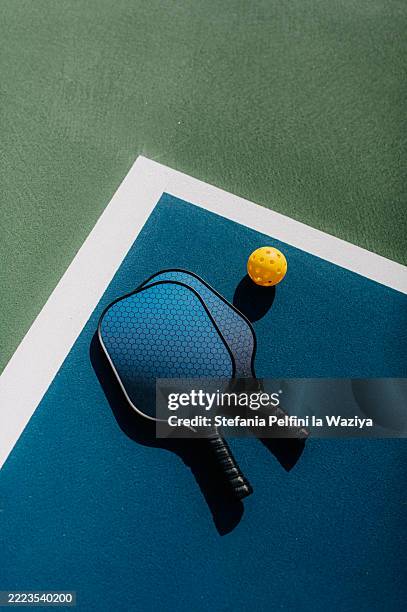 two blue pickleball paddles and a yellow perforated ball are placed on a multicolored court - paddle ball stock pictures, royalty-free photos & images