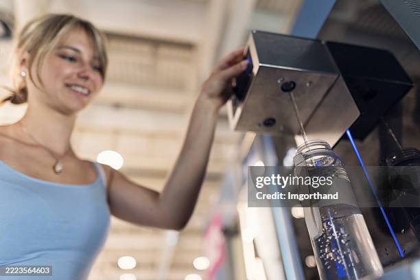 giovane donna felice che ricarica una bottiglia riutilizzabile da un distributore d'acqua automatizzato in un aeroporto. - ricaricare foto e immagini stock