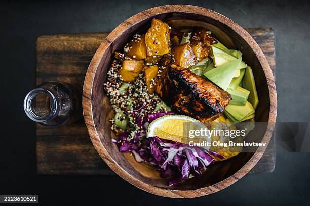 wooden bowl with fried salmon fish steak, quinoa, avocado, corn, cabbage salad and baked pumpkin. olive oil in bottle on side. dark black background, top view. healthy organic food concept. - quinoa stock pictures, royalty-free photos & images
