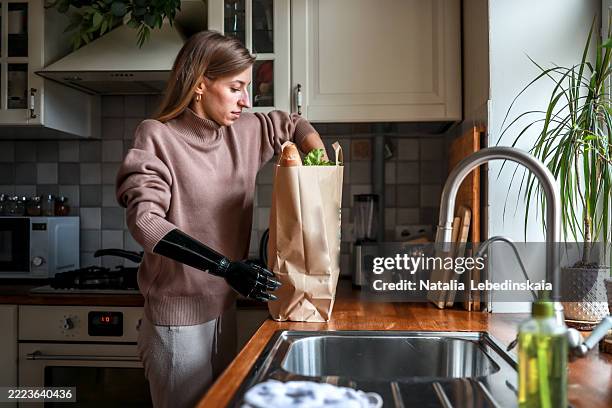 blonde woman with a prosthetic arm spends time in a cozy kitchen choosing fresh groceries from a large paper bag. - home delivery stock pictures, royalty-free photos & images