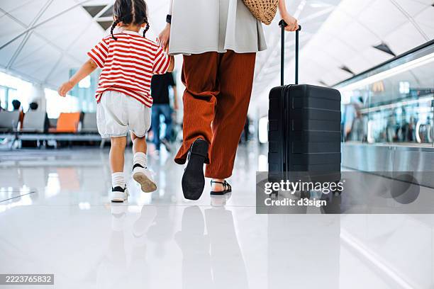 rear view of young asian mother carrying a suitcase while holding hands of her little daughter, walking through airport terminal and travelling by plane on a vacation. family travel and vacation. ready for a trip. embark on a new journey with the family - homecoming stock pictures, royalty-free photos & images