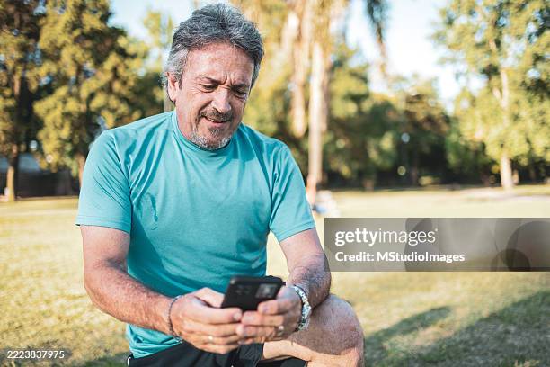 senior man checking his smart phone after exercising in the park - male likeness stock pictures, royalty-free photos & images