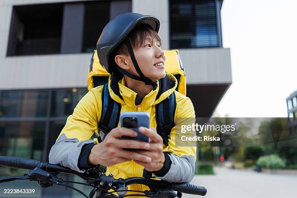 young deliveryman in a yellow jacket checking his phone while resting outdoors - side hustle stock pictures, royalty-free photos & images