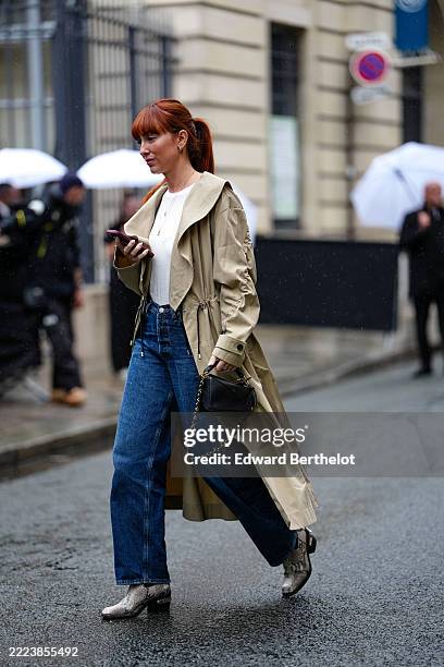 Guest wears a beige trench coat with a wide collar and belted cuffs over a white top. Blue denim jeans are paired with the outfit. Accessories...