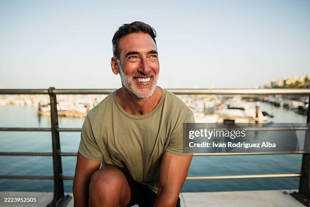 uomo maturo felice che sorride e si inginocchia in riva al mare - solo un uomo maturo foto e immagini stock