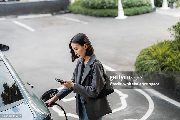 a woman leaning against her ev car and texting friends on her smartphone while enjoying a moment of relaxation at the charging station - convenience stock pictures, royalty-free photos & images