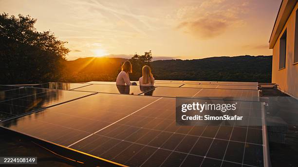 madre e hija en la instalación solar en la azotea viendo la puesta de sol dorada sobre las montañas - energía solar fotografías e imágenes de stock