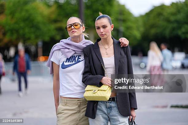 Model wears oversized amber-tinted sunglasses and a light purple sweater draped over the shoulders. The white T-shirt features a prominent blue logo...