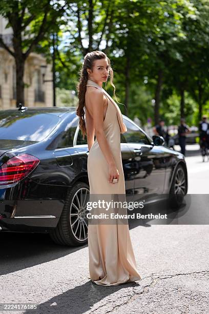 Guest wears a floor-length, sleeveless gown in a light beige hue, featuring a draped cowl neckline. The fabric appears smooth and slightly...
