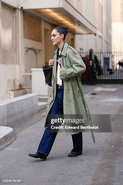 Model wears hair styled in a sleek bun, with bold eye makeup. An olive green trench coat features a classic double-breasted design with visible...