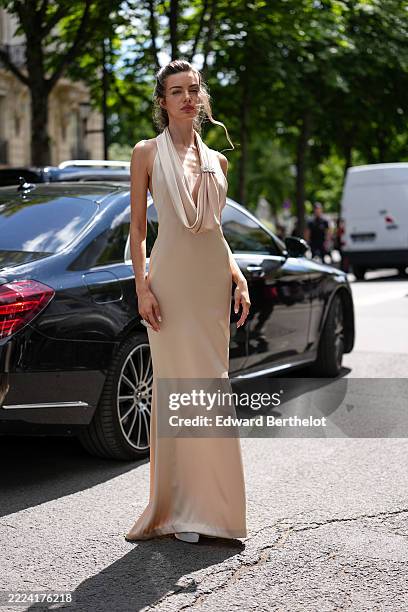 Guest wears a floor-length, sleeveless gown in a light beige hue, featuring a draped cowl neckline. The fabric appears smooth and slightly...