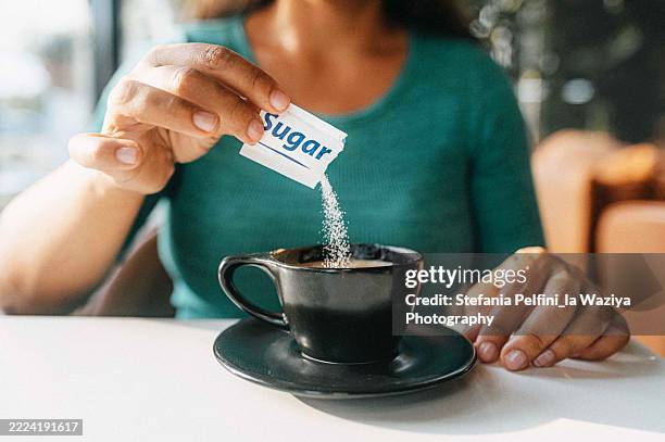 a woman’s hand pours a sugar packet into a black coffee cup, sitting at a bright, sunlit table. - sweetener packet stock pictures, royalty-free photos & images