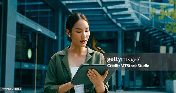 nahaufnahme einer jungen asiatischen geschäftsfrau, die vor einem modernen bürogebäude spazieren geht und digitale tablets und kabellose kopfhörer verwendet. remote-arbeit und nachhaltiges wirtschaften im freien. - sich verschönern stock-fotos und bilder