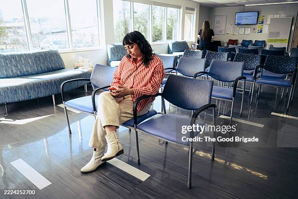 woman using smart phone in waiting room at medical clinic - benen over elkaar geslagen stockfoto's en -beelden