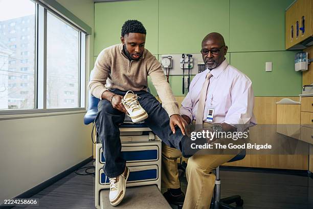 young man explaining ankle pain to senior male doctor - ortopedista imagens e fotografias de stock