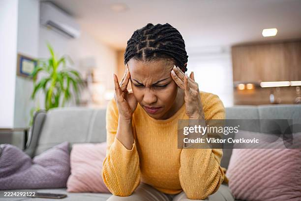junge frau, die unter kopfschmerzen leidet und ihre schläfen berührt, während sie auf dem sofa sitzt - laubsäge stock-fotos und bilder