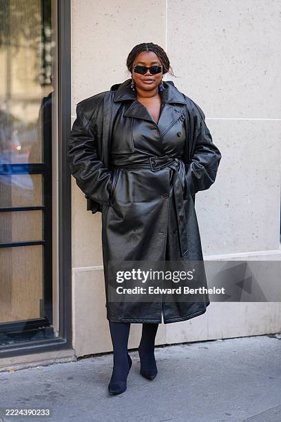 Guest wears black sunglasses, dark blue earrings, shiny black belted leather Balenciaga trench coat, black pointed toe Balenciaga heel tights,...
