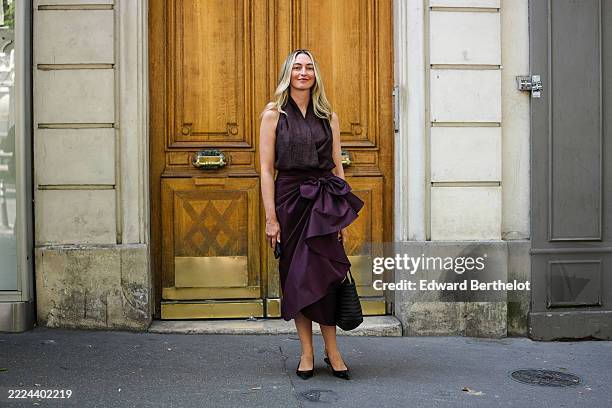 Guest wears a sleeveless, dark plum top with a subtle sheen, paired with a matching plum skirt featuring a large bow detail and cascading ruffles....