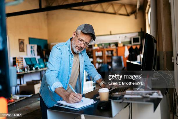mature entrepreneur writing notes and using computer in his workshop - klein bedrijf stockfoto's en -beelden