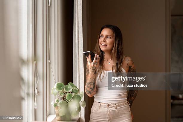 frau mit sprachassistent auf dem smartphone am fenster zu hause - ärmelloses oberteil stock-fotos und bilder