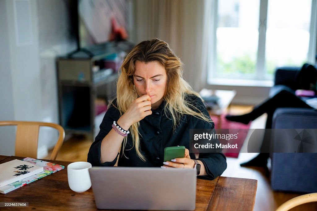 Worried female freelance worker scrolling smart phone while sitting at home office