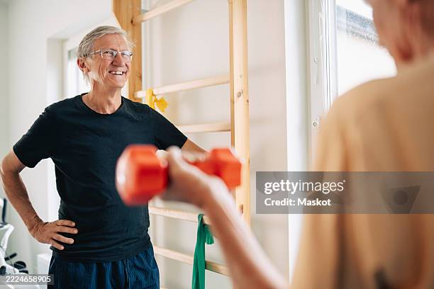 smiling elderly man standing with hand on hip while talking to male friend in rehab center - fitnessapparatuur stockfoto's en -beelden