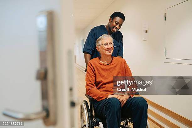 smiling male nurse pushing senior man sitting on wheelchair in alley at retirement home - duwen stockfoto's en -beelden
