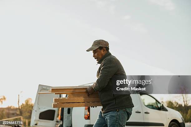 senior man carrying wooden planks while walking against sky at renovation site - bouwmateriaal stockfoto's en -beelden