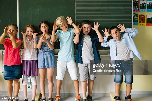 medium group of school children stand in front of on the classroom blackboard and making funny faces - summer school stock pictures, royalty-free photos & images