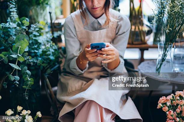 florist checking smartphone while surrounded by blooming flowers and greenery - florist bildbanksfoton och bilder