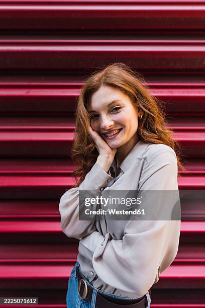 smiling redhead woman with hand on chin standing near corrugated iron - hand on chin stock pictures, royalty-free photos & images