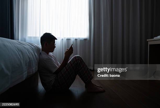 teenager boy sitting on the bedroom floor and using phone - depressie stockfoto's en -beelden