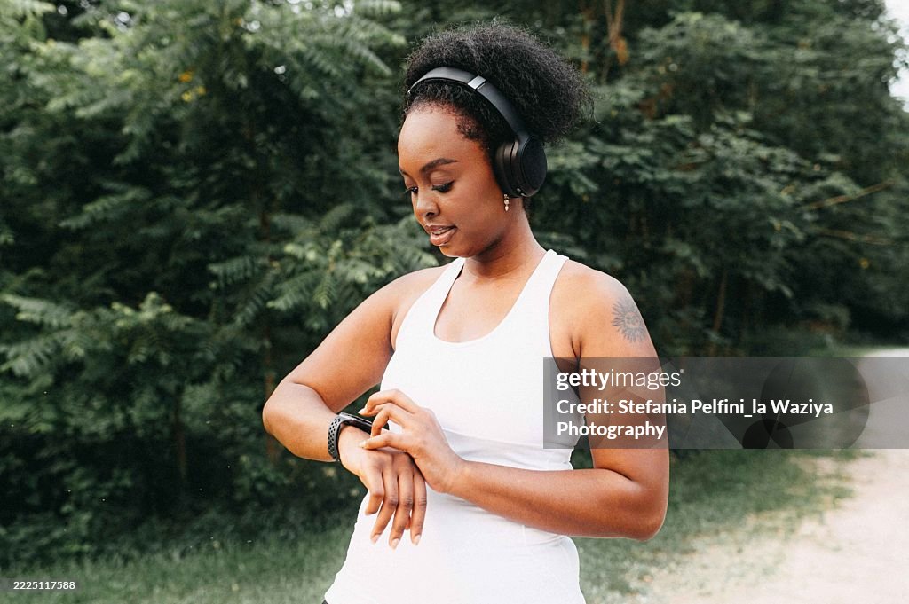 A woman runner stands on a path surrounded by lush greenery, checking her smartwatch while wearing wireless headphones.