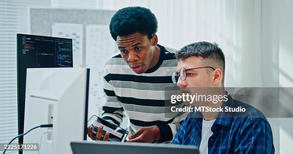 Two male software developers discussing project coding using digital tablet standing at desk in office. Artificial intelligence and programming. Two male software developers discussing project coding using digital tablet standing at desk in office. Artificial intelligence and programming.