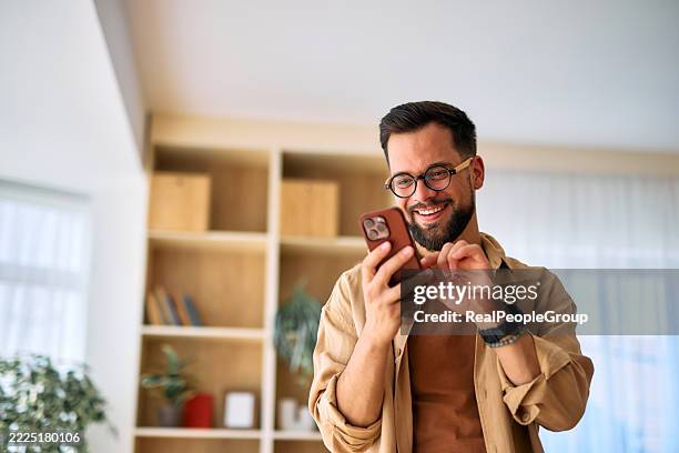 young man using smartphone at home, smiling and looking at screen - homens imagens e fotografias de stock
