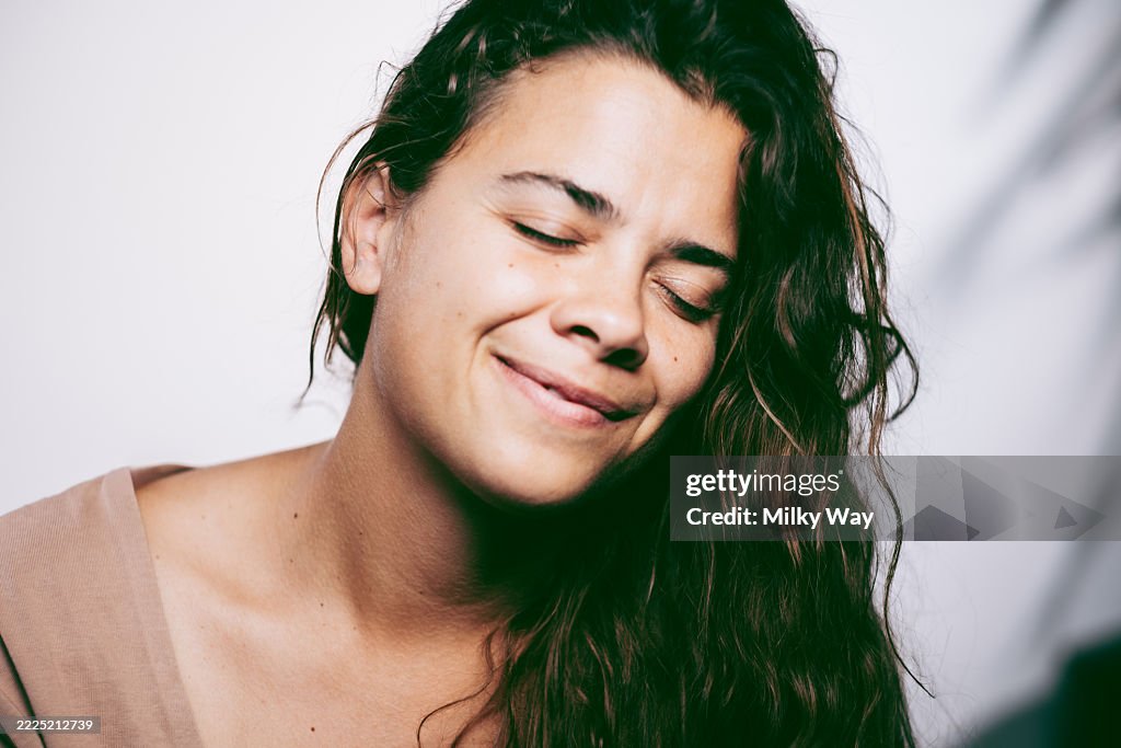 Young woman with long brown wavy hair smiles with eyes closed against plain background. Serenity, natural beauty and emotion concept.