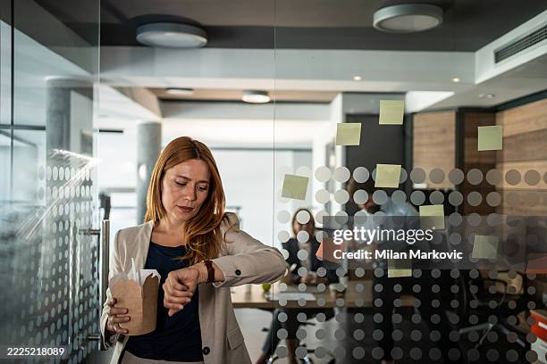 geschäftsfrau, die die zeit überprüft, während sie essen zum mitnehmen im büro hält - auf die uhr sehen stock-fotos und bilder