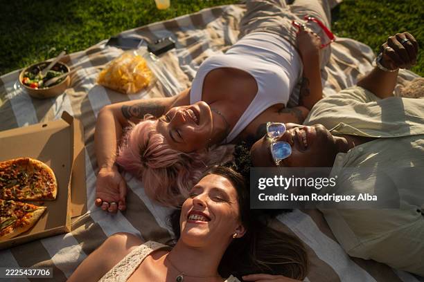 tres amigos acostados en una manta disfrutando de un picnic soleado en el parque - vida sencilla fotografías e imágenes de stock