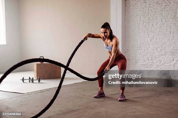 a focused and athletic young woman of fit build uses battle ropes during an intense workout in a minimalist gym. the bright setting highlights her dedication to fitness and strength training. - battle rope stock pictures, royalty-free photos & images