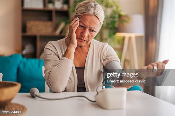 senior woman measuring blood pressure at home feeling sick - hypertensive stock pictures, royalty-free photos & images