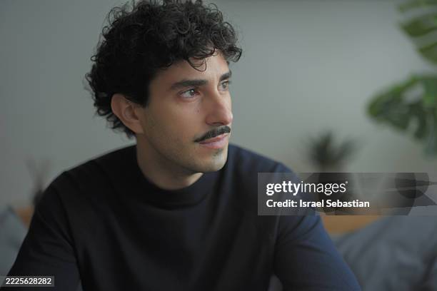 pensive young man with mustache looking away while sitting on sofa at home - mustache stock pictures, royalty-free photos & images