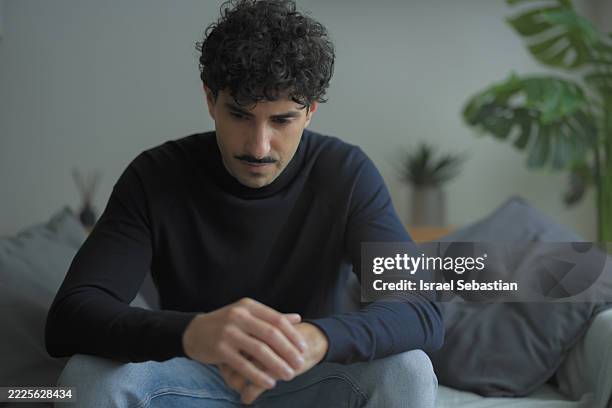 sad young man sitting on sofa at home reflecting on problems - southern europe stock pictures, royalty-free photos & images