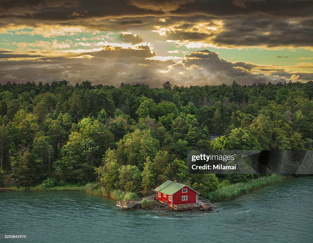 Aerial view of a red cabin, stormy sunset clouds, sun rays, forest and beach surrounded by beautiful sea views