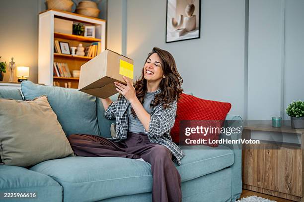 young woman opening package while sitting on couch at home - express delivery stock pictures, royalty-free photos & images