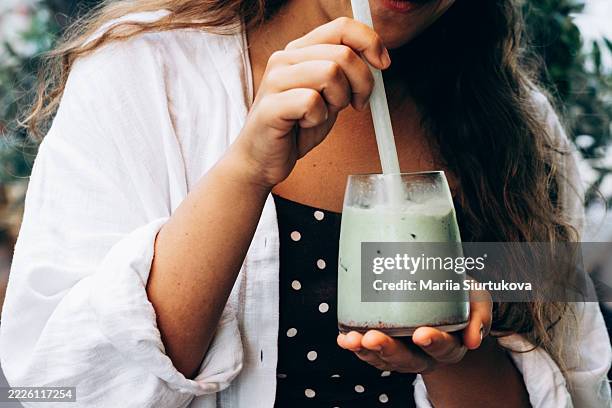 close-up of a woman's hand holding a glass of iced green matcha with straw. she wears a polka dot dress and white shirt. healthy lifestyle and refreshment. - woman adaptogen stock pictures, royalty-free photos & images