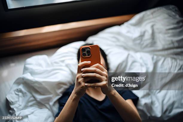 high angle shot of young asian woman texting on her smartphone while lying on bed relaxing at cozy home. chatting online with friends. social media. youth culture. lifestyle and technology - rencontre sur internet photos et images de collection