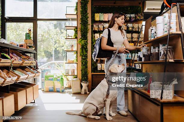 young woman shopping with her dog in a modern pet store, choosing pet supplies and dog food together. happy pet owner bonding with her dog a casual visit to the pet shop. - pet shop stock pictures, royalty-free photos & images