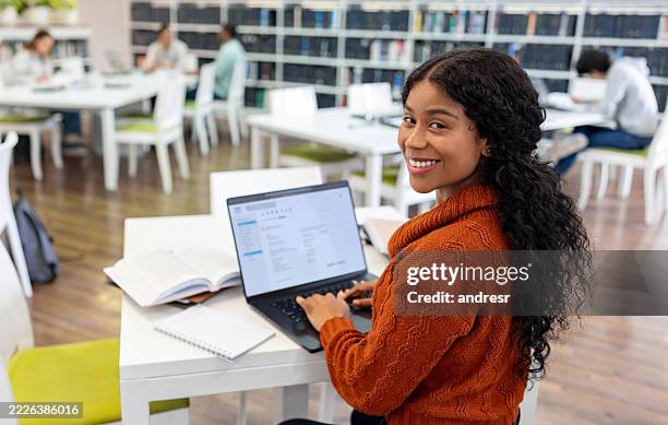 happy female student smiling at the library while using a laptop - public library stock pictures, royalty-free photos & images